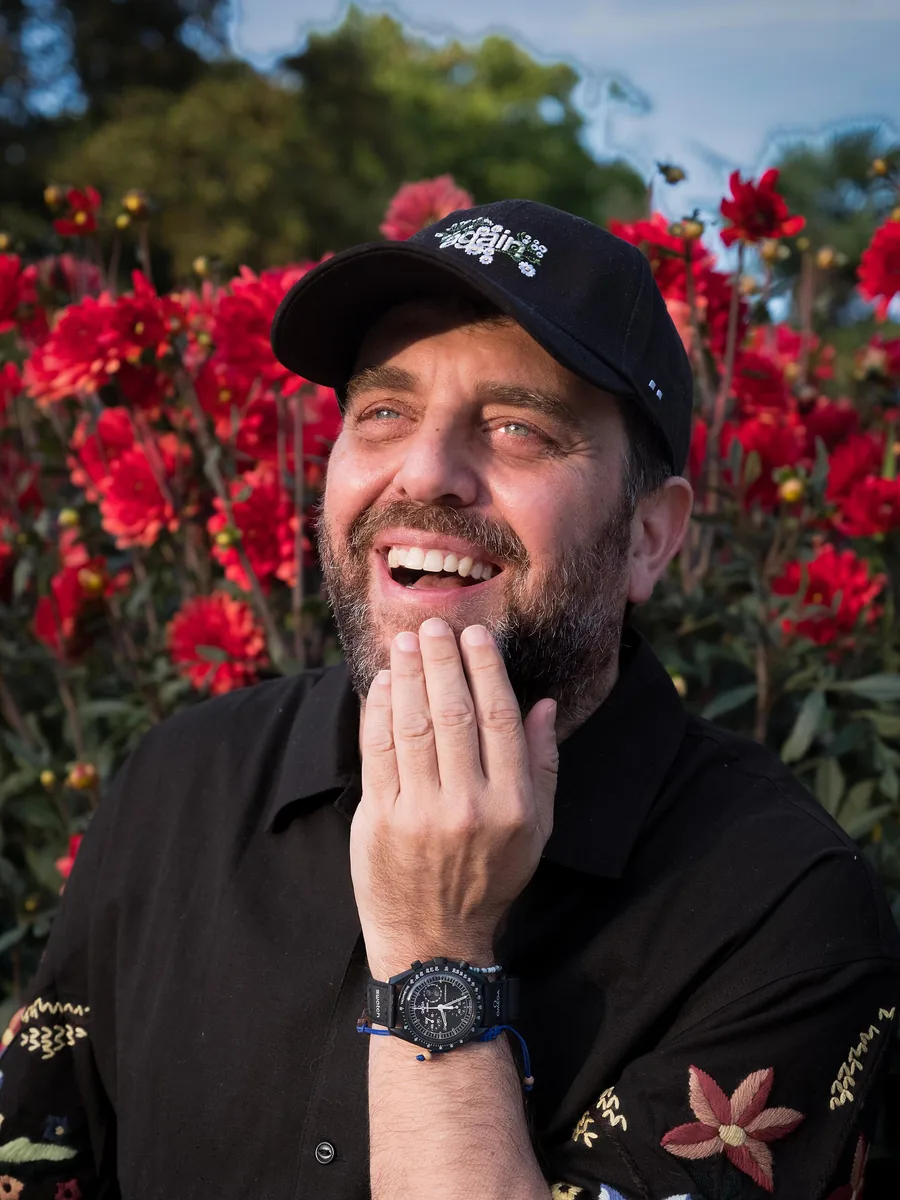 DJ Tennis sonriendo con gorra negra delante de unas flores rojas